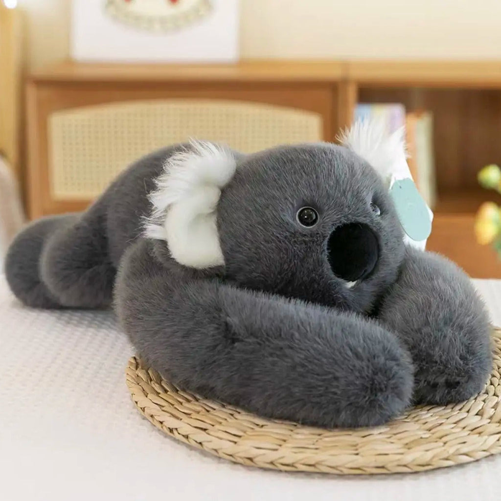 Gray koala plush toy with white ears and black nose, lying on a woven mat in a cozy indoor setting.