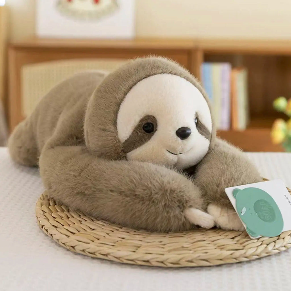 Soft brown sloth plush toy with white face and dark eye patches, lying on a woven mat in a cozy indoor setting.