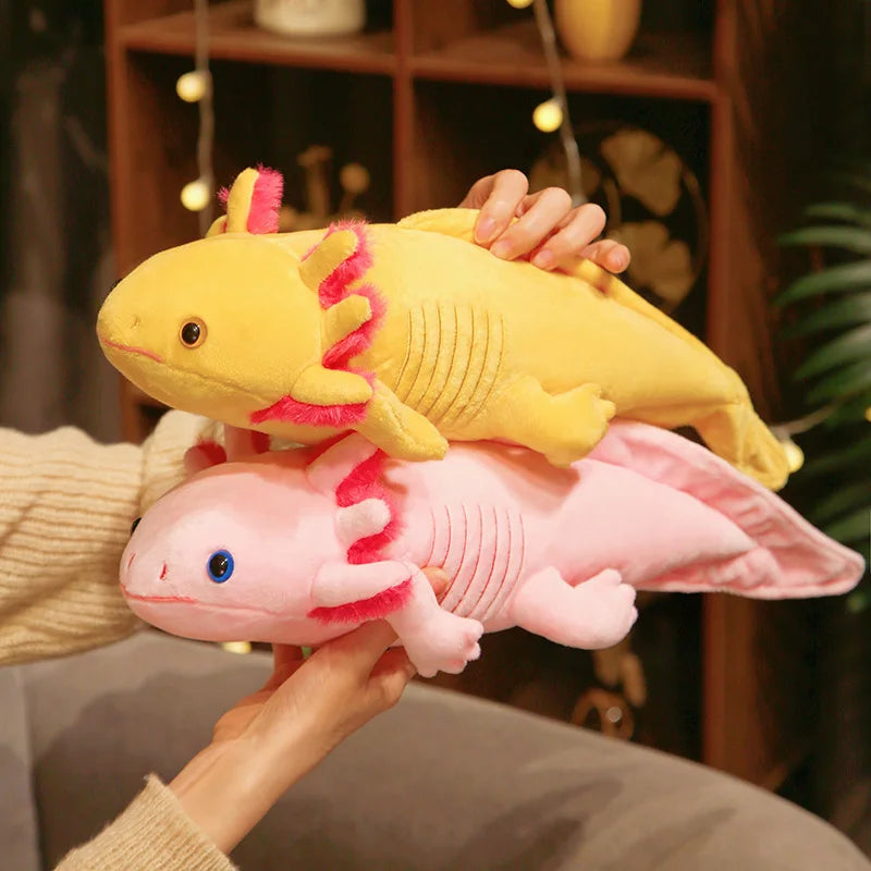 Two soft axolotl plush toys in pastel yellow and pink stacked together, featuring cute frilly gills and a cuddly long body design, held indoors for a cozy, playful look.
